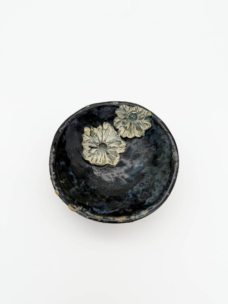 Adelina Ruvalcaba's ceramic bowl titled: "Fleeting Moments." A thrown bowl with dark glaze and two white flowers near the rim.